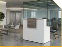 Modern office reception with a white counter, glass walls, gray chairs, and a potted plant on the left.