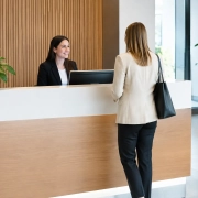 Cómo elegir mostradores de recepción para oficinas | OfficeDeco Receptionist in a black blazer smiles at a client standing at a white-and-wood front desk in a bright lobby with green plants nearby.