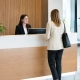 Cómo elegir mostradores de recepción para oficinas | OfficeDeco Receptionist in a black blazer smiles at a client standing at a white-and-wood front desk in a bright lobby with green plants nearby.